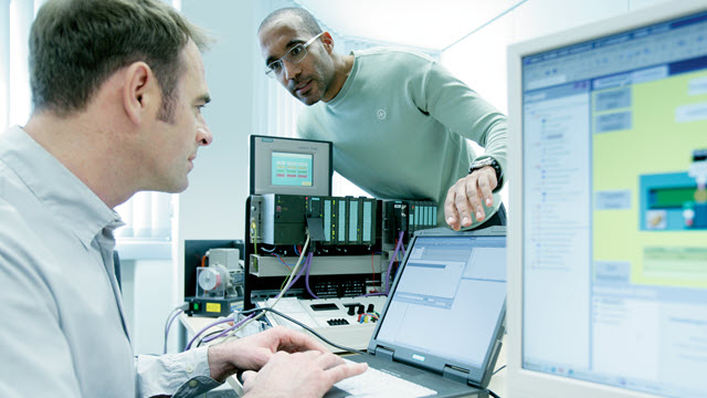 A man in an office is working on his laptop while another man leans over his desk to speak to him.