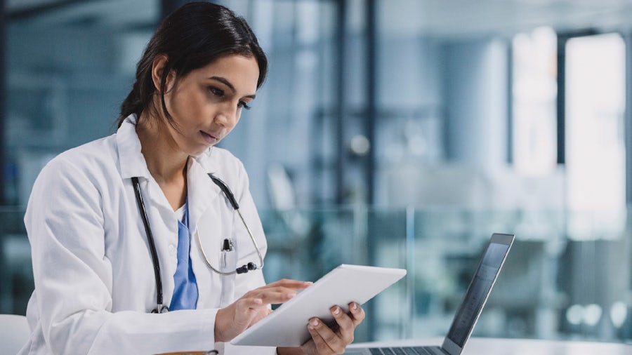 Female doctor sitting at a desk with a laptop while holding a tablet