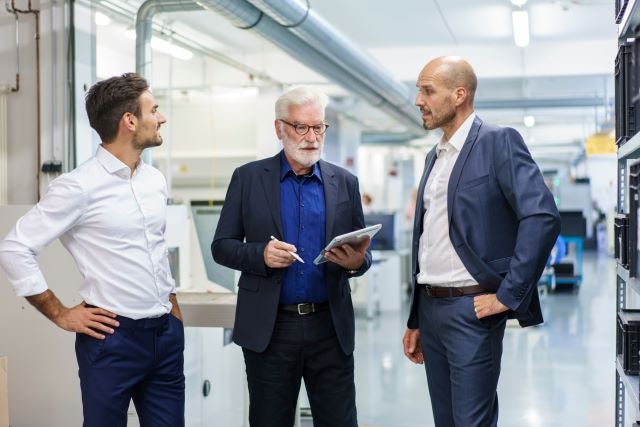 Three professionals discussing quality management in a manufacturing facility, with one holding a clipboard, indicating an inspection or evaluation.