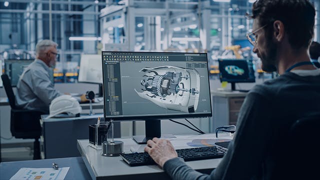 Engineer working on a 3D model prototype at a desktop computer in a robotic arm assembly line manufacturing facility.