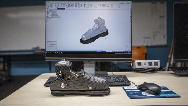 Computer screen with a digital mock-up of a prosthetic foot, and a real version of the prosthetic sitting on the desk.