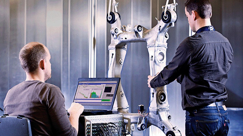 Dwóch mężczyzn pracujących nad częścią do maszyny. Jedna osoba korzysta z oprogramowania Simcenter do symulacji i testów na laptopie, podczas gdy druga przeprowadza inspekcję części.