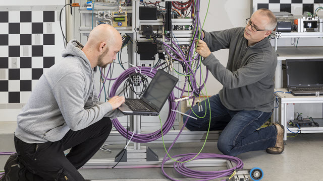 Two men in hoodies and work pants work with wiring and a laptop.