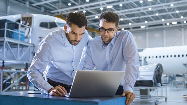 Dwóch mężczyzn w niebieskich koszulach przeglądających instrukcje pracy na laptopie.