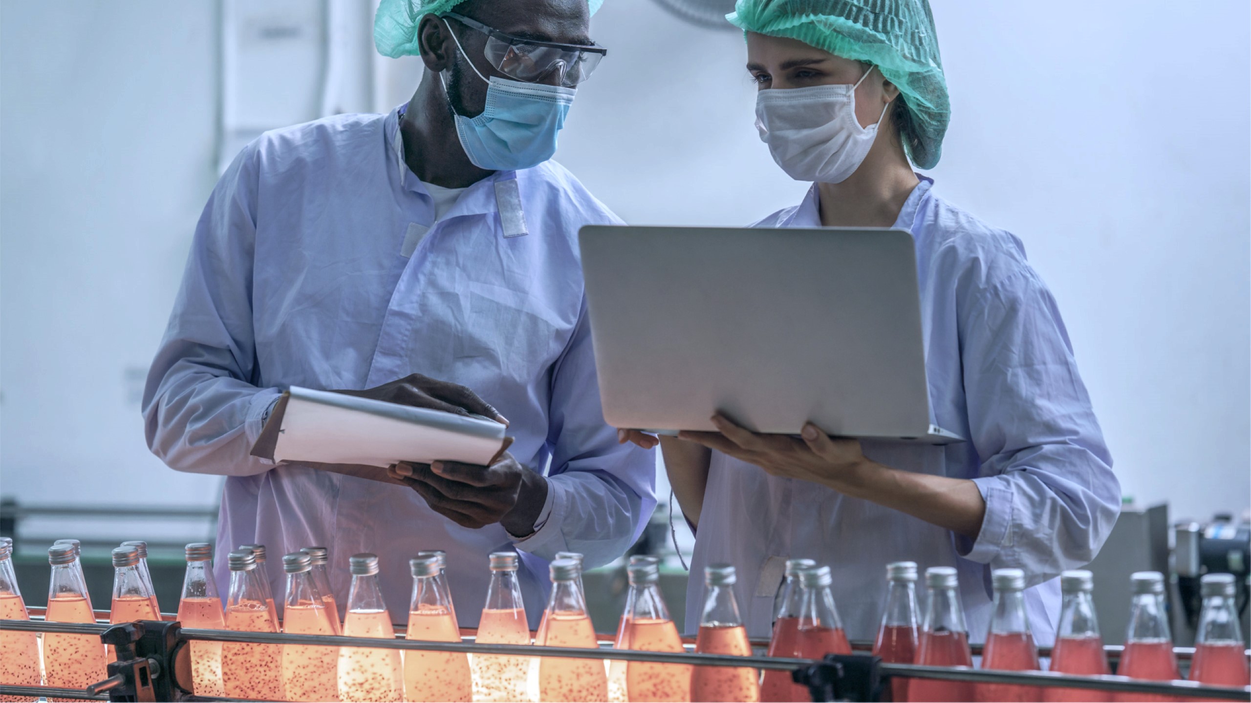 Two factory workers wearing PPE. One is holding a clipboard and the other is holding a laptop. They are examining bottled beverages on a factory line.