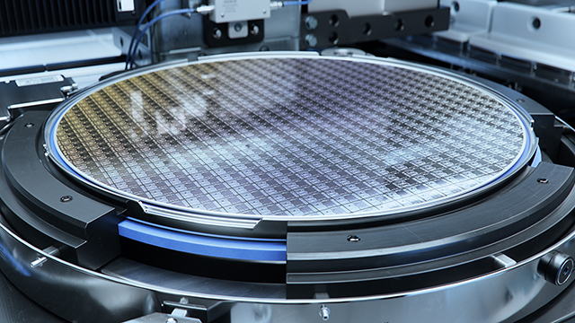 Close-up photo of a silicon wafer mounted in a circular testing or manufacturing platform, showing detailed grid pattern across the wafer's surface with metallic and blue mounting hardware around the edges.
