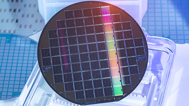 Silicon wafer in a clear plastic holder showing a dark surface with grid pattern of rectangular sections. A vertical stripe of rainbow colors (green, orange, pink) runs through the center of the wafer.
