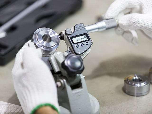 Two gloved hands use a quality gauge to measure a machine part