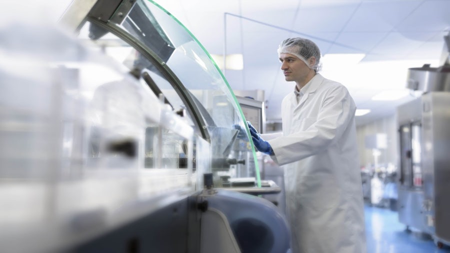 A person in a lab coat, hairnet, and gloves observes machinery with a transparent shield in a cleanroom manufacturing environment.