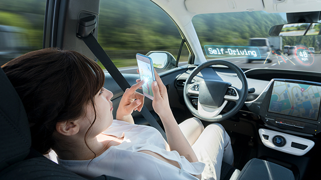 A woman relaxes in the front passenger seat of a self-driving car, using her smartphone while the vehicle autonomously navigates the road with digital speed and navigation displays.