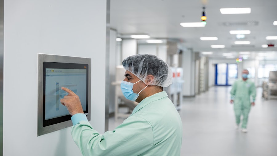 Man in cleanroom suit and mask operating a wall-mounted touchscreen in a pharmaceutical manufacturing plant.