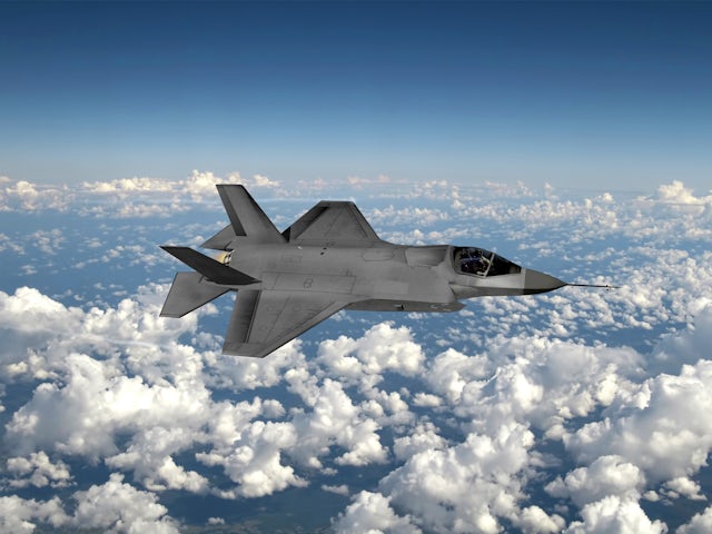 A miliary aircraft soars in the sky, above a layer of scattered clouds.