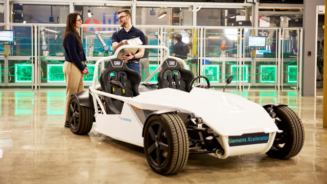 The image shows a white, open-top two-seater vehicle labeled "Siemens Xcelerator", parked indoors on a shiny floor. In the background, two people are conversing in a tech-focused setting with equipment and screens.