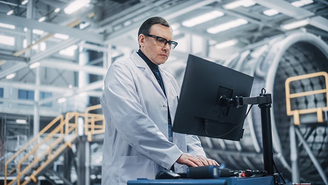 A worker is wearing a white lab coat and standing in front of a computer monitor viewing the screen. They are in a large-scale manufacturing facility.