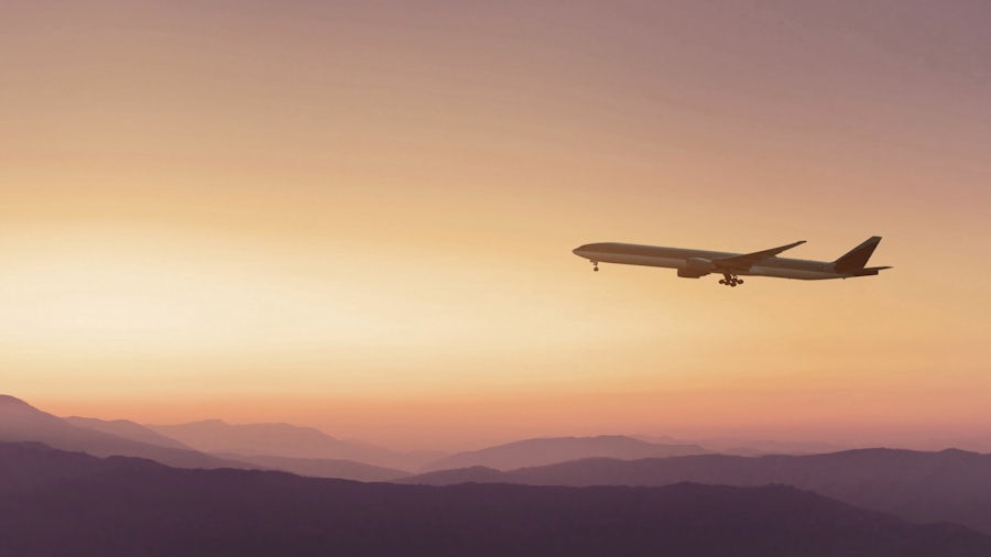 A plane flies over mountains as the sun sets in the background