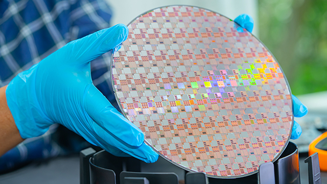 Hands wearing blue nitrile gloves hold a silicon wafer showing a checkerboard pattern of integrated circuits with rainbow-like light diffraction effects visible across its surface.