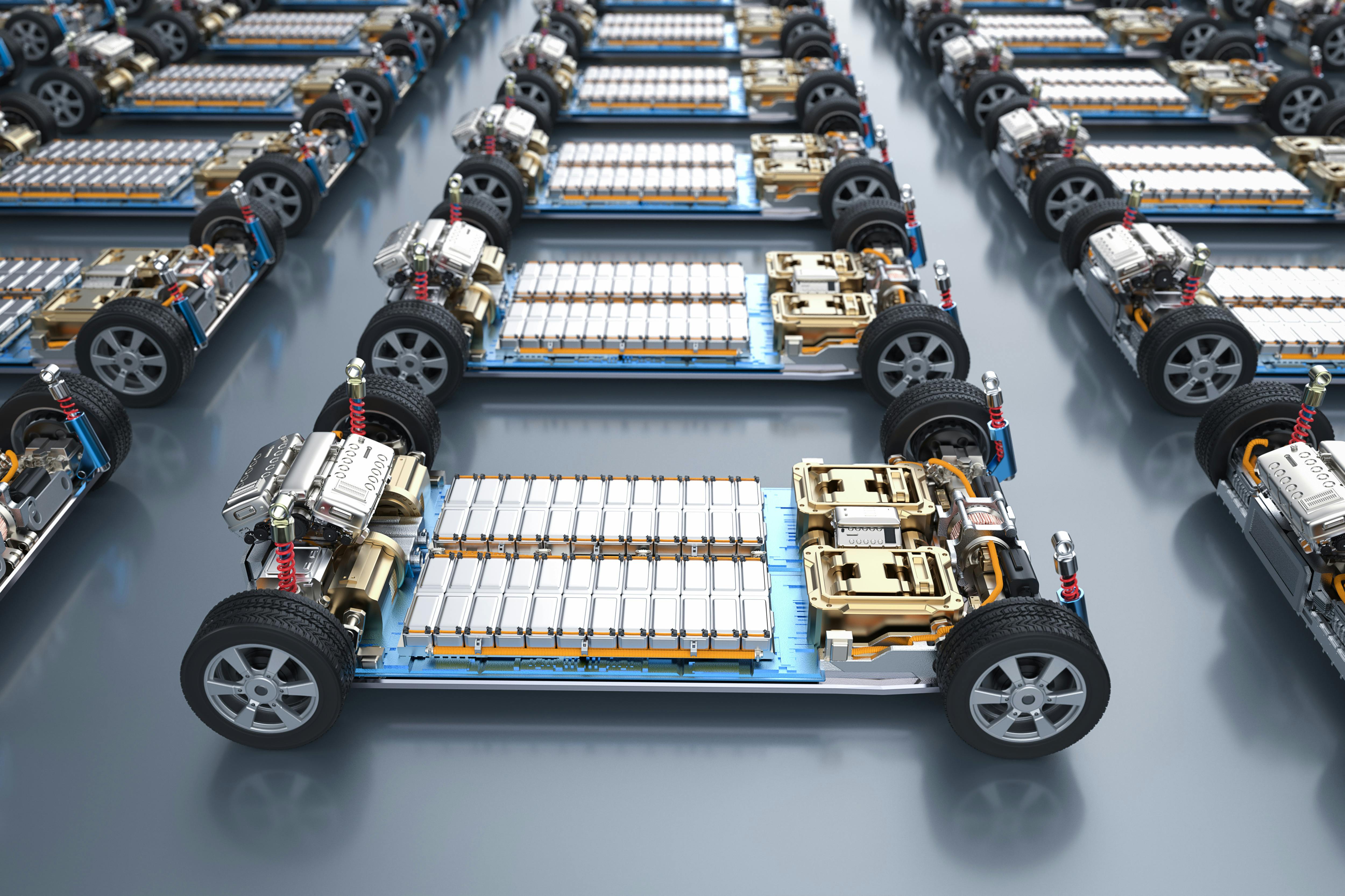 The chassis of an electric car packed with battery cells. The chassis includes twin motors and the four tires. There is one EV in the foreground surrounded by dozens of others.