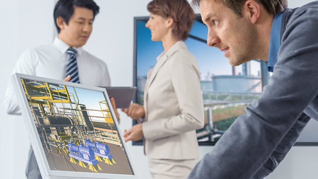 A man is studying his laptop monitor. In the background, a woman and a man in business attire are conversing.