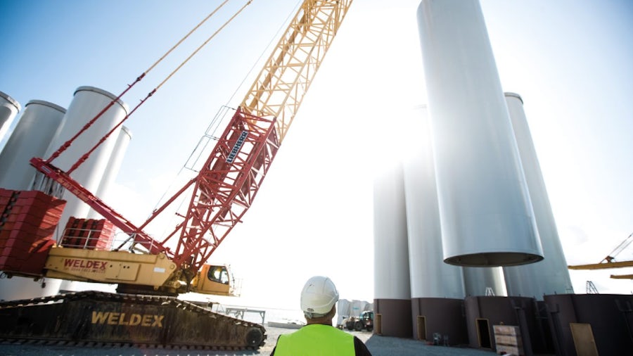A man in a white hard hat and bright yellow vest watches as a crane lifts parts at a sustainable energy plant.