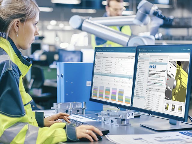 A woman in a factory environment is wearing a yellow and black jacket in front of a computer screen.