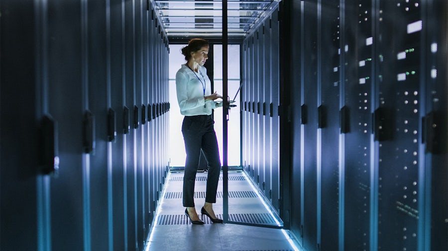 A woman in a white shirt and dark pants stands in a server room, holding a laptop. Rows of server racks with blue lighting line the aisle, and the floor also has illuminated strips.