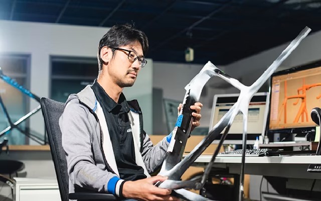 Man with glasses holding a Giant-brand metal bike frame with a design of the bike frame within Designcenter NX CAD software in the background