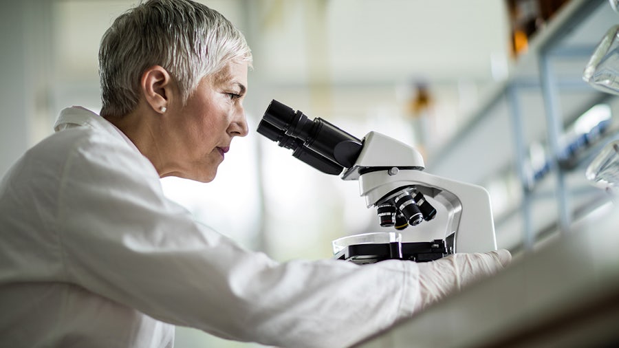 Scientist wearing a white lab coat looks through microscope.