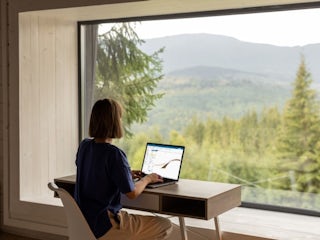 A woman at her desk facing a window, where there is a forest and hills outside. She is using her laptop with <a href=