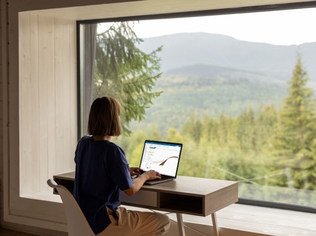 A woman at her desk facing a window, where there is a forest and hills outside. She is using her laptop with an NX CAD window open.