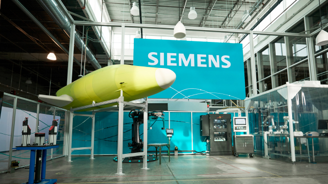 Industrial facility with a large yellow-green object suspended from the ceiling, a blue SIEMENS banner in the background, and various equipment including computers and enclosed workstations.
