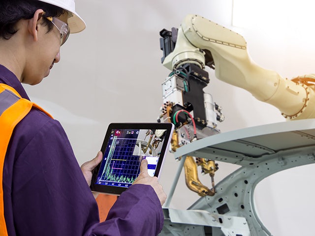 A man wearing a reflective vest, goggles, and a hard hat looks at a tablet with a robot's performance. There is a robot arm blurred in the background.