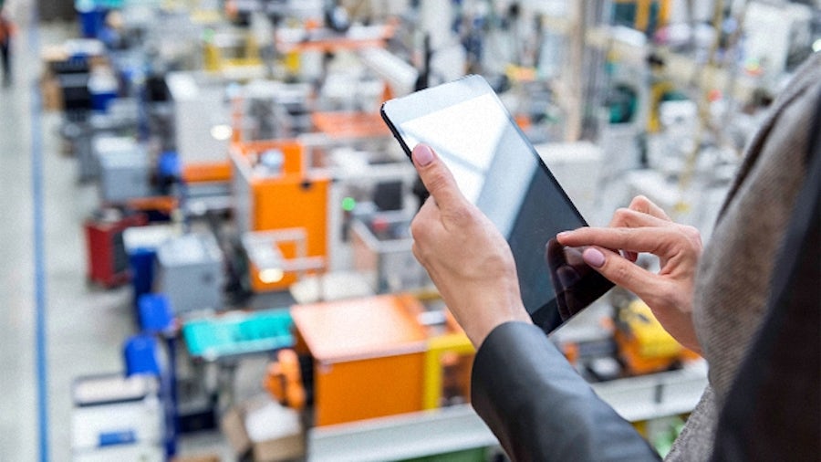 A person holding a tablet and using manufacturing operations management software like Opcenter to oversee the manufacturing floor below.