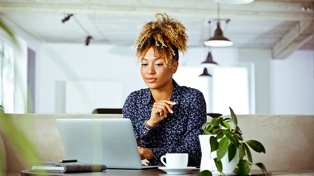 A woman at a table in a cafe with a coffee. She is reading a blog article from the laptop in front of her.