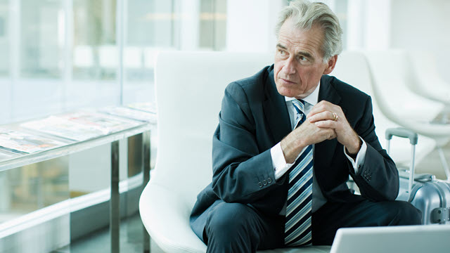 A man in a suit is seated in an office chair in front of a laptop with hands clasped.
