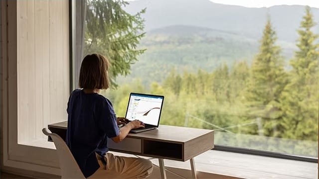 A woman sitting at a desk overlooking a forest. Her hands are on the keyboard of a laptop.