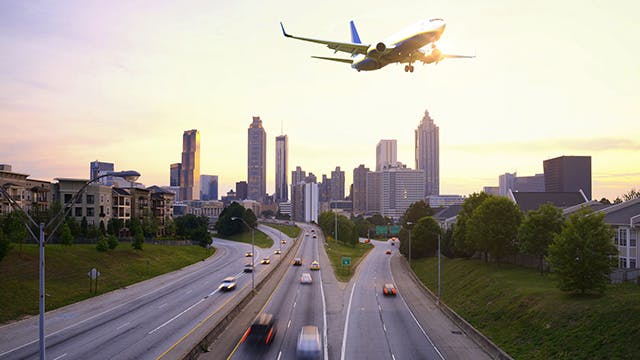 An airplane flies over several lanes of a highway, between treelined areas of residential buildings, with a city skyline in the background.