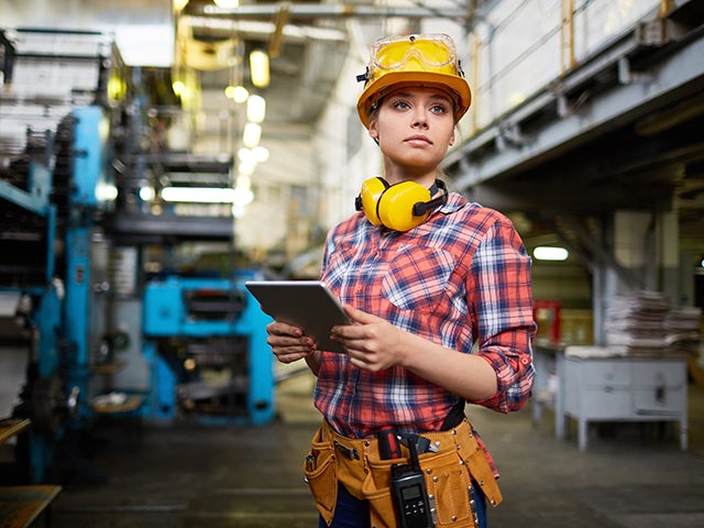 A production planning and scheduling worker standing on the shop floor holding a tablet. Wearing a hard hat and protective headphones.