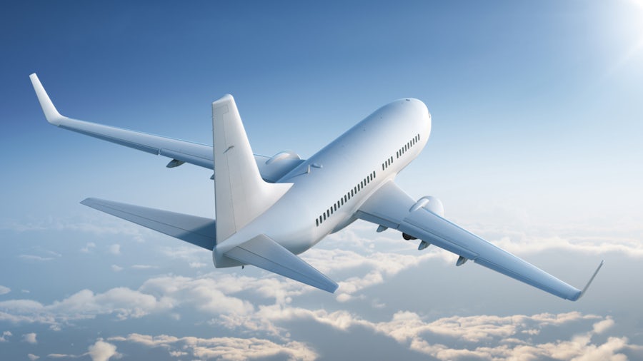 An all-white airplane flies toward the sun with a light blue sky and fluffy clouds in the background