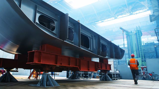 Portion of ship hull structure in construction on supports with a man in protective gear walking past.