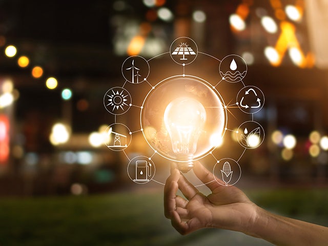 A hand holding up a glowing light bulb with luminous icons around it, implicating different environmentally-friendly ways of generating energy.