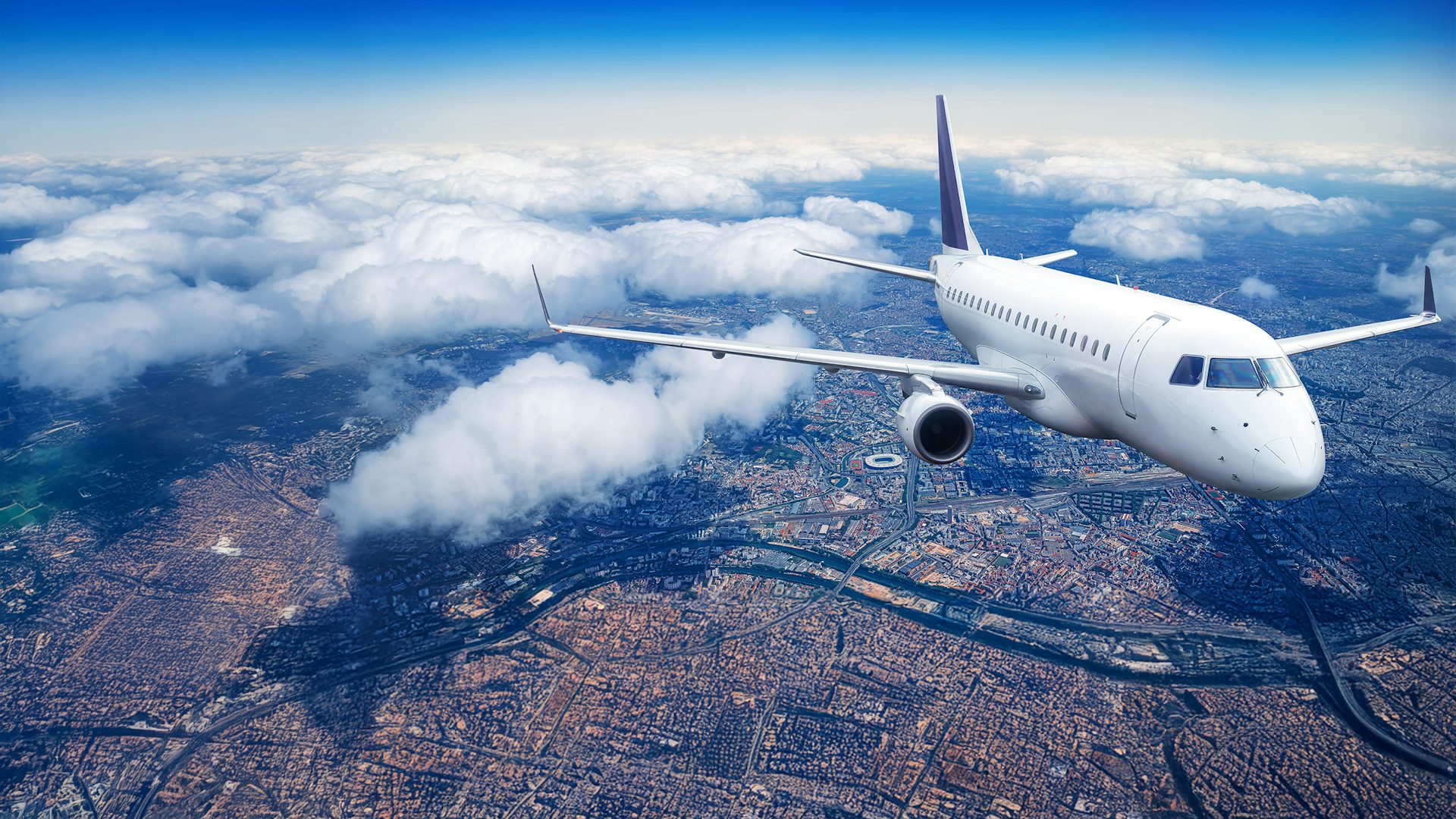 Commercial airplane flying above a cityscape, surrounded by clouds and blue sky.