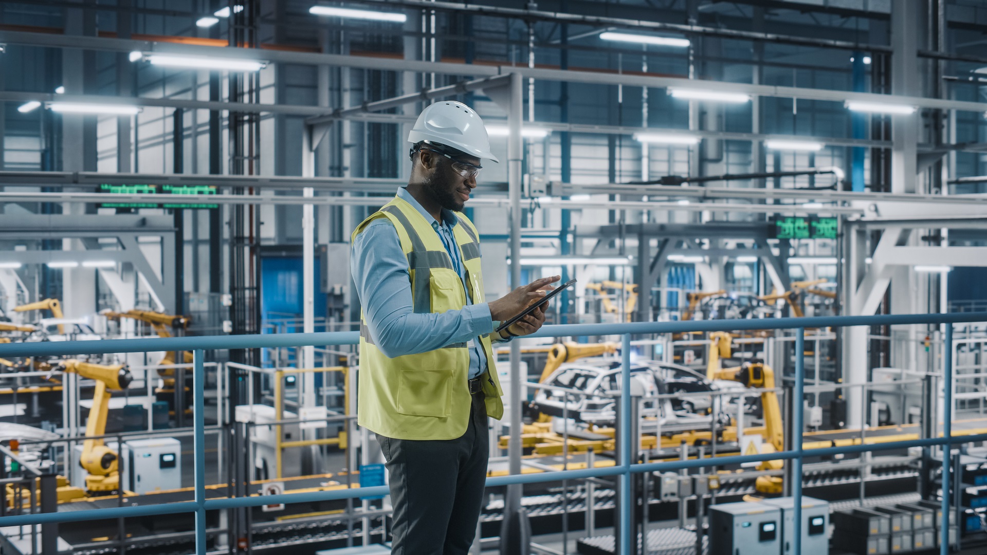 A man in a hard hat looks at a tablet with a factory in the background
