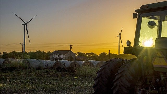 Un tractor en una granja con molinos de viento y una puesta de sol de fondo.