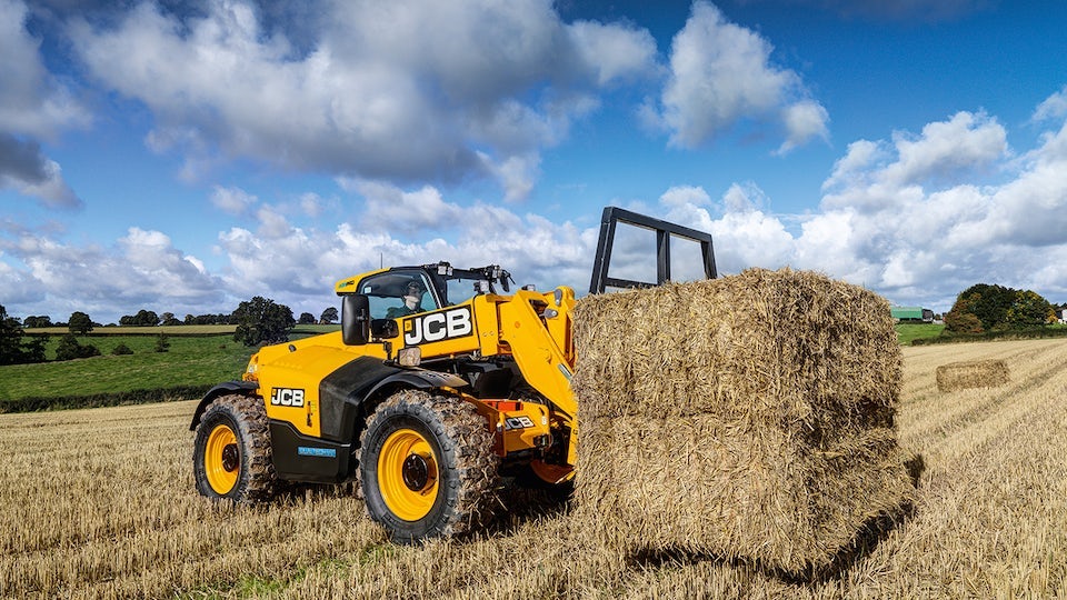 A JCB forklift lifting a bale of hay in a field.