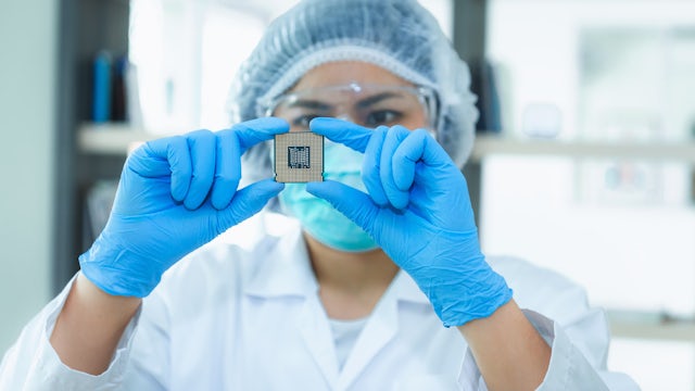 A technician wearing a hairnet and goggles inspects a semiconductor