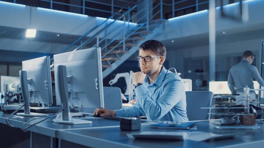 Electronics development engineer sitting at his desk uses personal computer for programming.