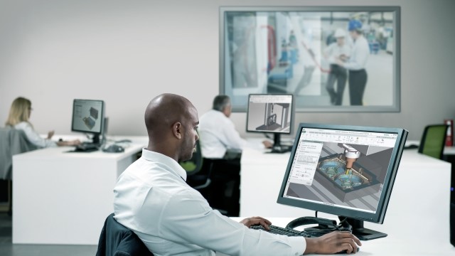Three engineers at their desks in the office of a manufacturing facility. Each engineer has an instance of NX for manufacturing software on their desktop screen.