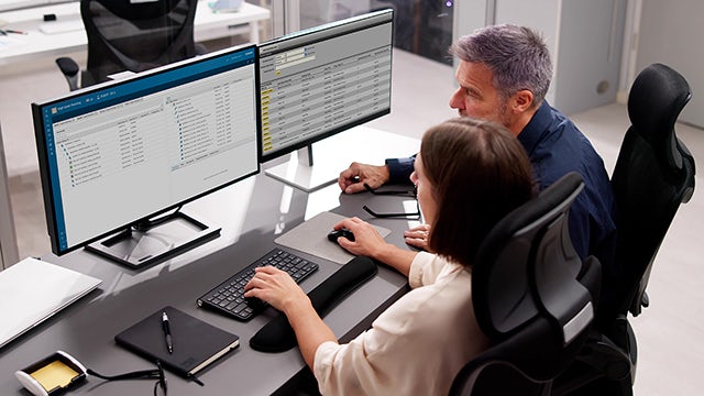 Un homme et une femme assis l'un à côté de l'autre à un bureau, regardant deux écrans d'ordinateur dans un environnement de bureau.