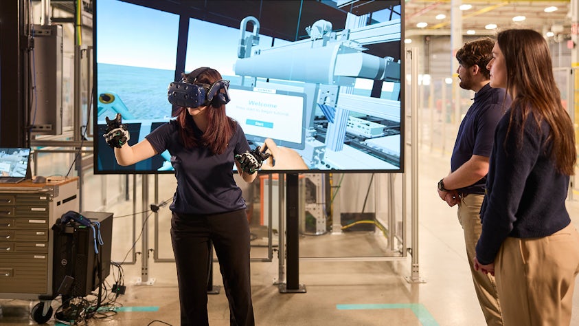 A woman is engaged with a virtual reality headset and gloves, interacting with a screen that displays an industrial or mechanical setting. Two others are watching the experience, suggesting a tech demonstration or training session.