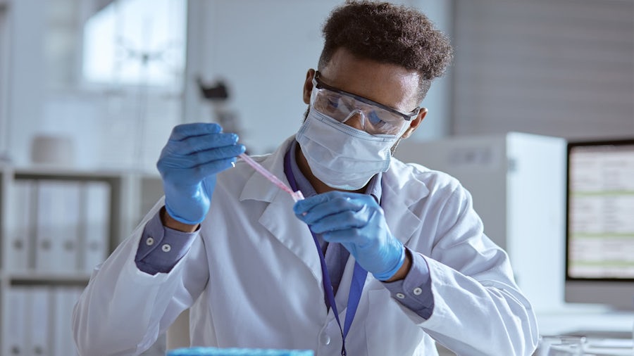 A male researcher wearing a lab coat, face mask, safety goggles, and blue gloves, precisely pipetting a red liquid into a small test tube in a laboratory setting.